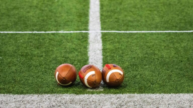 Três bolas de futebol americano alinhadas sobre um campo de grama artificial verde. Elas estão posicionadas lado a lado, bem em cima de uma linha branca central do campo, com outra linha branca horizontal ao fundo. A imagem destaca a textura da grama e o formato oval das bolas, sugerindo um cenário esportivo antes do início de um jogo ou treino.
