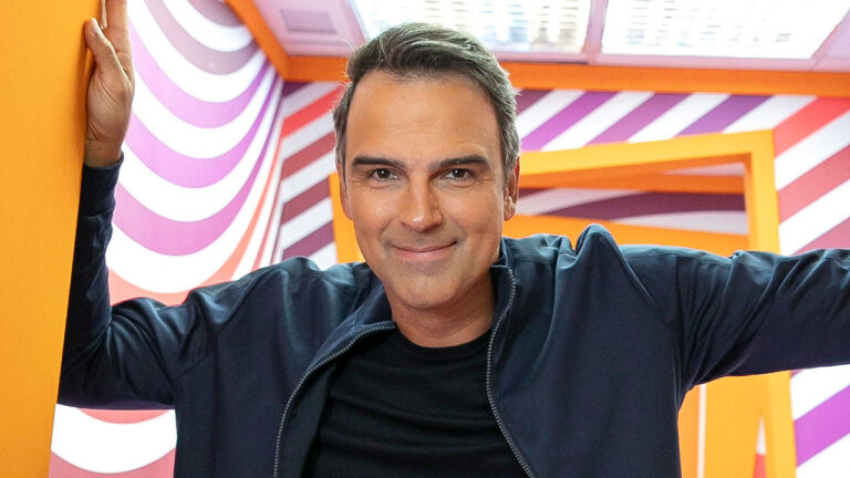 Tadeu Schmidt sorri no cenário colorido e vibrante do Big Brother Brasil com paredes de listras roxas e laranjas na casa da Globo