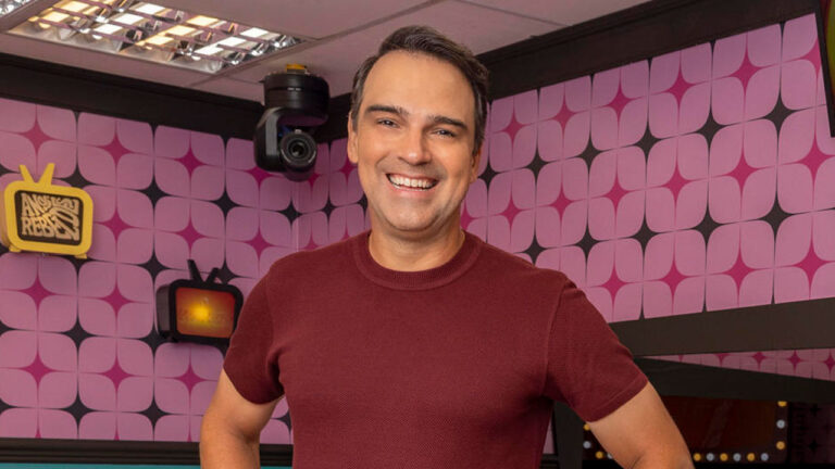 Tadeu Schmidt sorri no cenário do Big Brother Brasil com paredes decoradas por padrões geométricos rosa e roxo nos Estúdios Globo