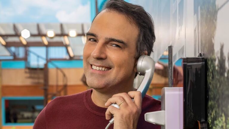A imagem mostra Tadeu Schmidt sorrindo, usando suéter vermelho e segurando um telefone fixo prateado. Ele está em um ambiente colorido e iluminado, na casa do BBB, transmitindo uma atmosfera descontraída e comunicativa