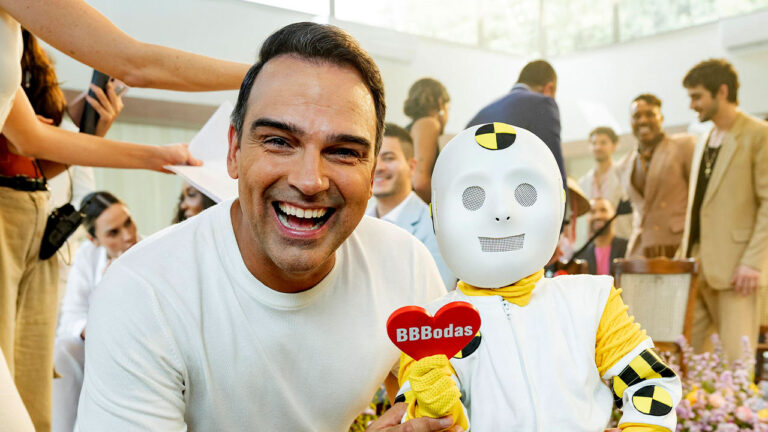 Tadeu Schmidt sorri ao lado de Dummynho que segura um coração escrito BBBodas durante gravação do Big Brother Brasil