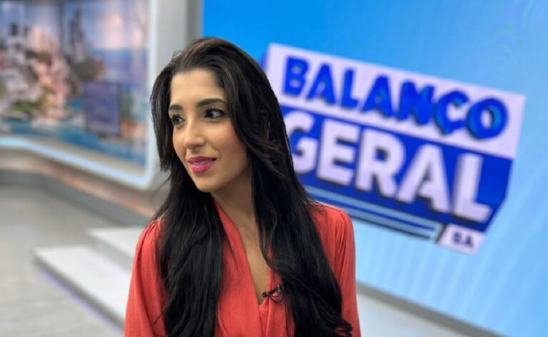 Uma apresentadora de televisão aparece em estúdio, em plano médio, vestindo uma blusa vermelha de mangas longas. Ela olha levemente para o lado, com expressão serena. Ao fundo, um telão azul exibe o logotipo “Balanço Geral”. O cenário é um estúdio de telejornal com iluminação clara e elementos gráficos modernos.