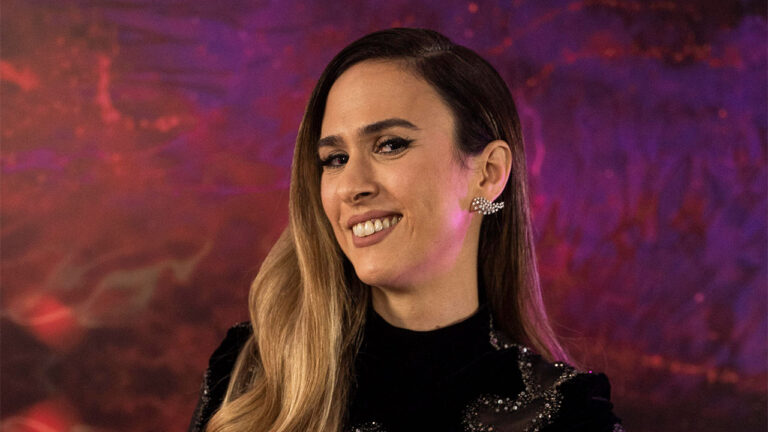 Tatá Werneck sorri usando vestido preto com bordados prateados e brincos brilhantes em frente a um fundo abstrato roxo e laranja