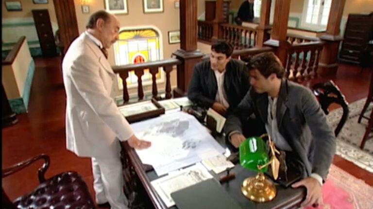 Cena em um escritório antigo, com mobiliário de madeira escura. Um homem mais velho, de terno claro, está de pé ao lado da mesa mostrando um grande mapa aberto para dois homens sentados, que o observam atentamente. A iluminação é quente e há vitrais coloridos ao fundo