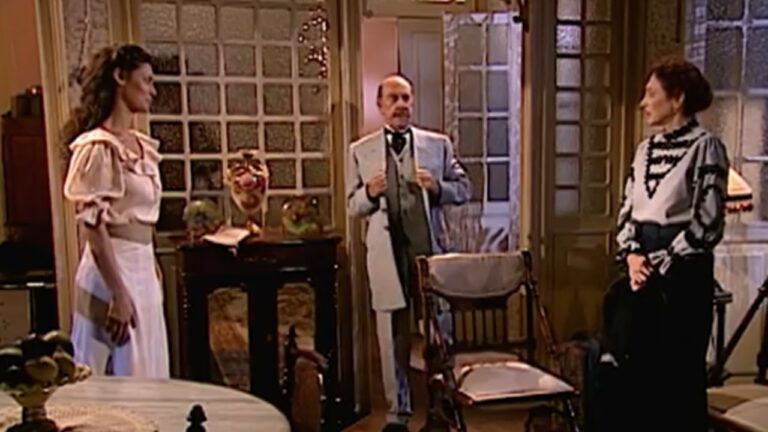 Cena de época em ambiente interno, com três pessoas em uma sala bem iluminada. À esquerda, uma mulher jovem usa vestido claro com avental, em pé e voltada para o centro do cômodo. No meio, um homem mais velho, de bigode, veste terno claro e segura a lapela do casaco, posicionado próximo a uma cadeira de madeira. À direita, uma mulher mais velha, sentada, usa roupa escura com detalhes claros e observa os outros personagens. O cenário tem portas de vidro, paredes com blocos translúcidos e móveis clássicos, sugerindo uma residência antiga.