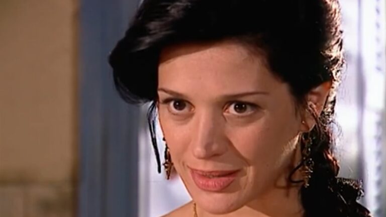 Close no rosto de uma mulher adulta de cabelos escuros presos, com mechas soltas na lateral. Ela usa brincos delicados e olha diretamente à frente, com expressão intensa e levemente irônica. A iluminação é quente e o fundo desfocado indica um ambiente interno, sugerindo uma cena dramática de novela ou série.