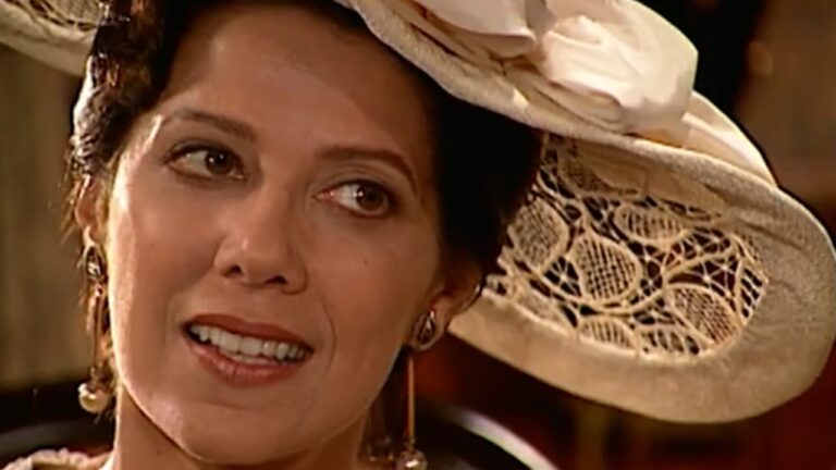 Uma mulher aparece em close, em ambiente interno com iluminação quente. Ela usa um chapéu claro de estilo antigo, com renda delicada e detalhe floral, inclinado levemente para o lado. Tem expressão suave e atenta, com um leve sorriso, olhando para alguém fora de quadro. Usa brincos discretos e maquiagem natural. A composição, o figurino e a iluminação sugerem uma cena de época, possivelmente de filme ou novela ambientada no início do século XX.