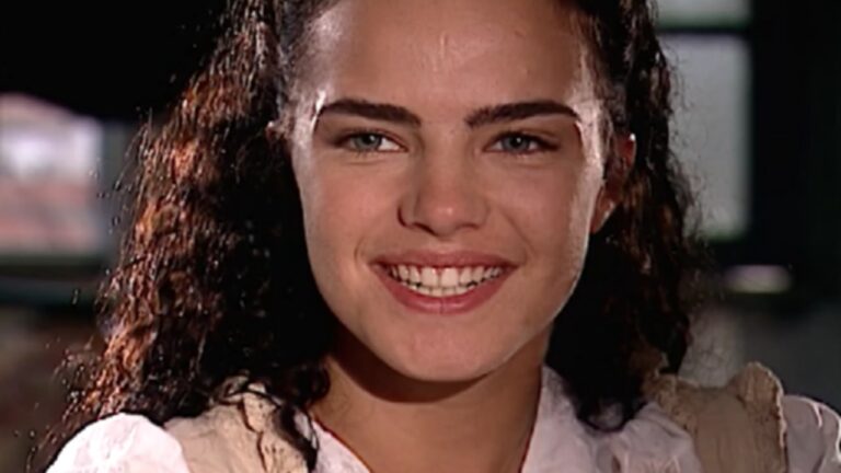 Mulher de cabelos cacheados sorrindo em cena de época com blusa clara e iluminação suave ao fundo