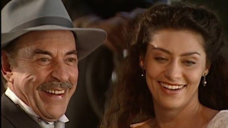 Homem mais velho, usando chapéu cinza e terno, sorri amplamente ao lado de uma mulher jovem de cabelos longos e escuros, também sorridente. Ambos aparecem em um ambiente interno com luz quente, transmitindo clima de alegria e cumplicidade