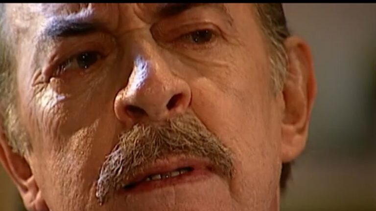 Close extremo do rosto de um homem mais velho, com expressão séria e olhos marejados. O enquadramento destaca rugas, bigode espesso e a tensão em seu olhar, sugerindo um momento de forte emoção ou conflito.