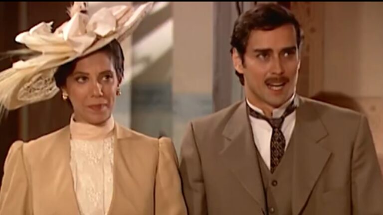 Homem e mulher vestidos com roupas de época, aparentando o início do século XX, estão lado a lado em um ambiente interno. A mulher usa um tailleur claro com gola alta rendada e um chapéu ornamentado com plumas; o homem veste terno bege, camisa branca e gravata, exibindo expressão falante enquanto a acompanha.