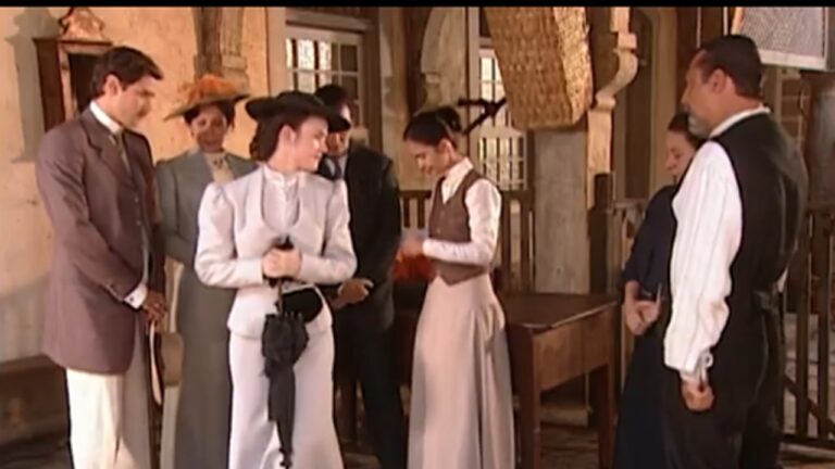 Grupo de pessoas em ambiente interno com arquitetura antiga, vestindo roupas de época claras e escuras; algumas mulheres usam chapéus elegantes e seguram acessórios, enquanto conversam e se organizam próximas a uma mesa de madeira, sugerindo uma cena histórica ou de novela de época.