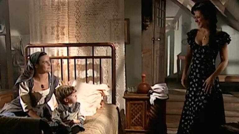 Em um quarto simples com decoração rústica, uma mulher sentada na beira da cama, usando vestido e lenço na cabeça, observa ao lado de um menino pequeno que também está sentado. Diante deles, outra mulher em pé, vestindo um vestido longo estampado, sorri calorosamente. A luz suave entra pela janela com cortina rendada, reforçando o clima acolhedor e doméstico da cena