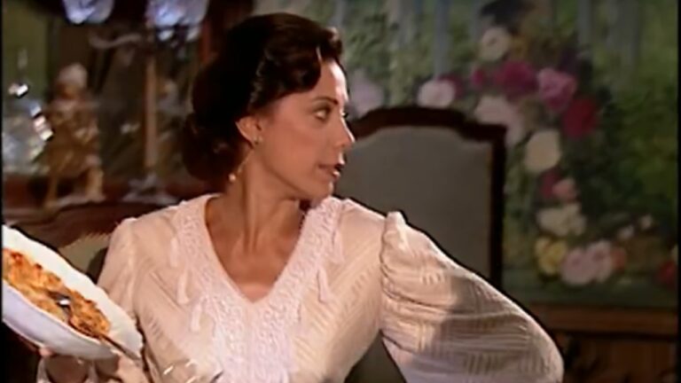 Mulher com vestido branco de época segura prato de comida e olha para o lado em cena ambientada em sala decorada com papel floral