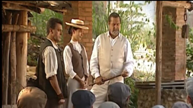 Homem de colete claro e camisa branca segura um bastão enquanto fala diante de um grupo de pessoas sentadas, acompanhado por uma mulher de chapéu e colete marrom e outro homem de colete escuro, em ambiente rústico com pilares de madeira e tijolos aparentes