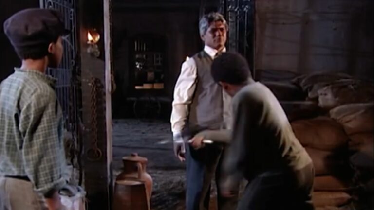 Cena em um armazém pouco iluminado, com sacos empilhados ao fundo. Um homem adulto está parado enquanto um jovem faz um movimento brusco em sua direção segurando um objeto metálico, sugerindo tensão ou confronto. Outro garoto observa a cena da entrada, próximo a uma porta de ferro.