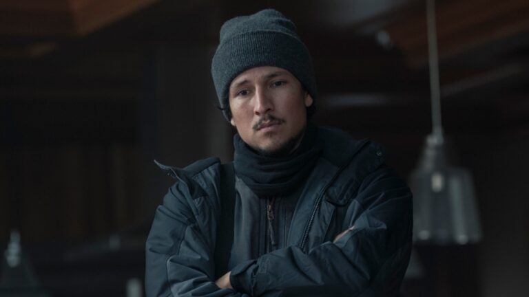 Homem com expressão séria veste gorro de lã cinza, cachecol preto e casaco grosso escuro, com os braços cruzados, em ambiente interno pouco iluminado com luminárias metálicas desfocadas ao fundo