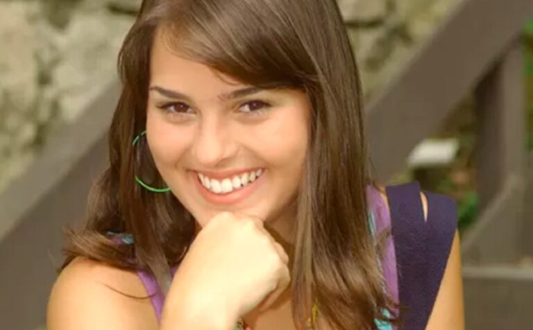 Mulher jovem de cabelos castanhos longos sorri com a mão apoiada no queixo, usando brincos grandes e coloridos. Ela está ao ar livre, com fundo desfocado que inclui vegetação e estruturas de madeira.