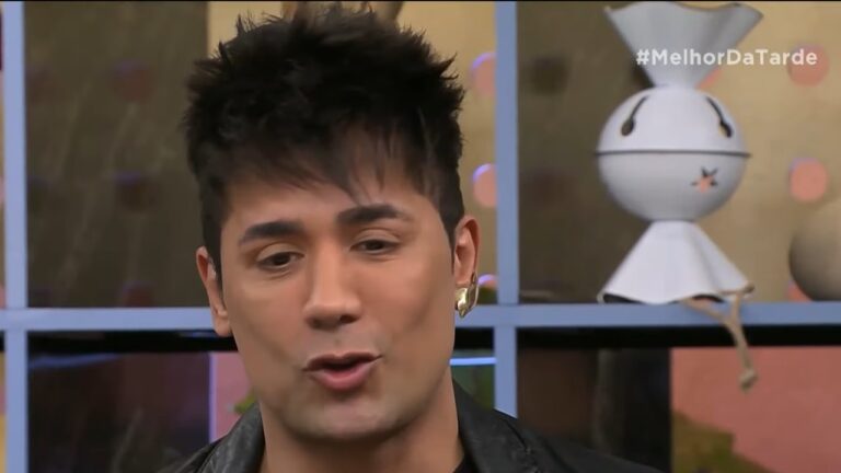 Homem com jaqueta preta e brinco dourado fala em estúdio do programa Melhor da Tarde com prateleira decorada ao fundo