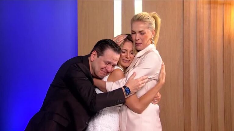 Três pessoas se abraçam emocionadas, com expressões de choro. Um homem de terno preto está à esquerda, e duas mulheres — uma de vestido branco e outra de blazer claro — estão à direita, em um ambiente interno com parede de madeira e iluminação suave.