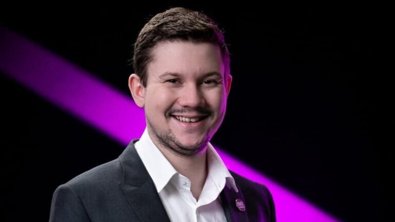 Homem jovem posa sorrindo em estúdio, usando terno escuro e camisa branca. O fundo preto é iluminado por um feixe diagonal de luz rosa, criando contraste e destaque para o retrato.