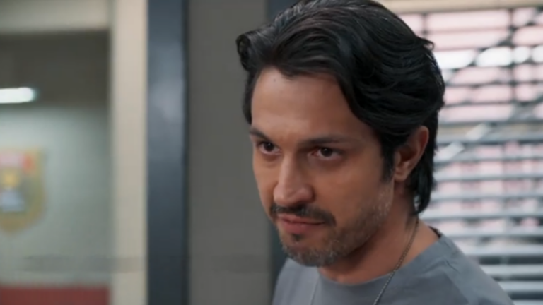 Homem aparece em plano fechado dentro de um ambiente interno, com expressão séria e olhar concentrado. Ele tem cabelo escuro penteado para trás e barba por fazer, veste camiseta cinza e usa um colar discreto. Ao fundo, há uma parede clara, uma porta de vidro com um símbolo desfocado e persianas que indicam um ambiente institucional, como escritório ou delegacia.