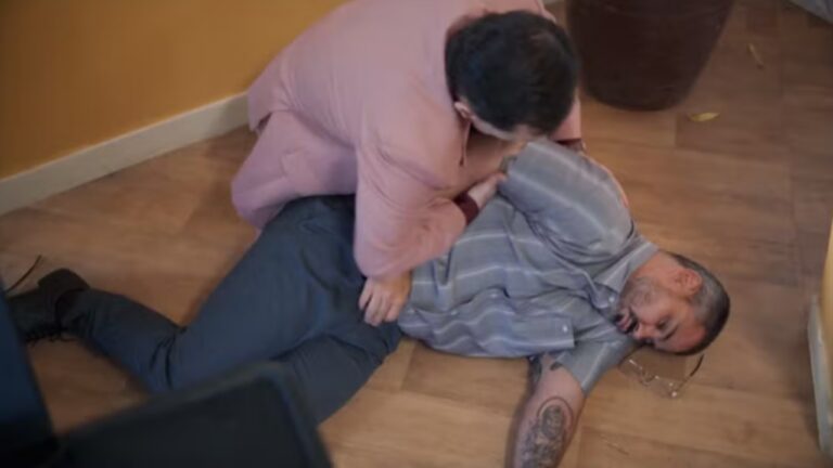 Homem de camisa cinza desmaiado no chão é socorrido por outro ajoelhado ao seu lado em cena de novela
