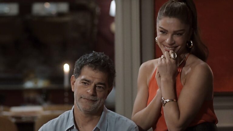 Homem de cabelos grisalhos e barba rala veste camisa azul clara e está sentado ao lado de uma mulher de cabelos presos em rabo de cavalo alto, usando blusa vermelha sem alças, brincos e anéis dourados, ambos sorrindo em ambiente interno iluminado por luz suave com vela acesa ao fundo