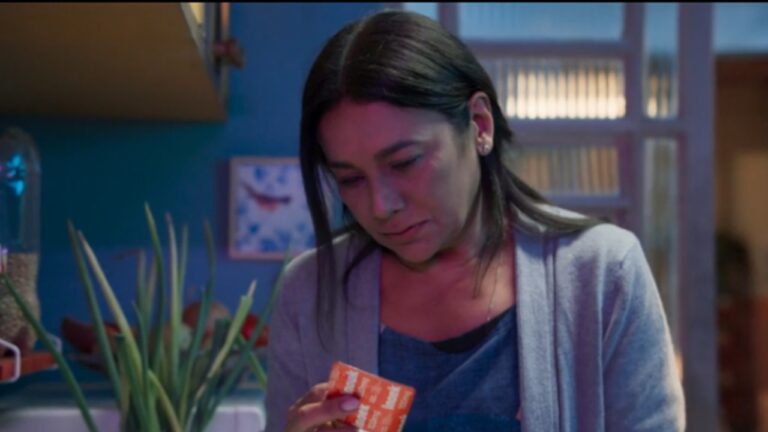 Mulher em ambiente doméstico pouco iluminado olha para uma caixa de medicamento laranja com expressão triste e cansada. Ela veste cardigã cinza sobre blusa azul, e ao fundo há plantas e objetos de cozinha desfocados, compondo um clima de preocupação ou reflexão.