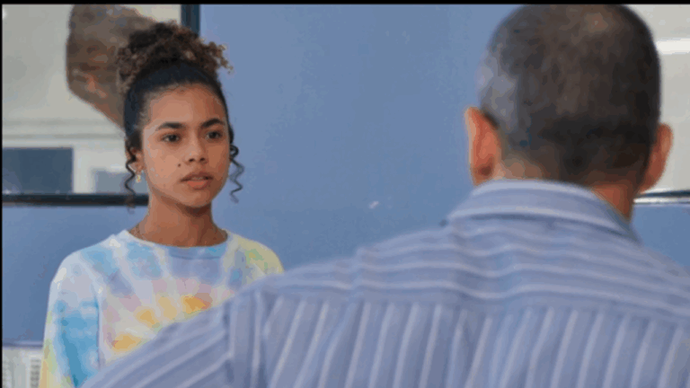 Jovem de cabelo cacheado preso em um coque alto olha séria para um homem de costas. Ela veste uma blusa colorida em tie-dye, enquanto o homem usa uma camisa listrada azul. Ambos estão em um ambiente interno de paredes azuis.