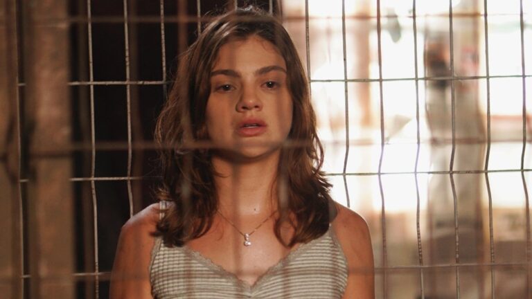 Mulher de expressão tensa e roupa simples está atrás de grade metálica em cena de novela