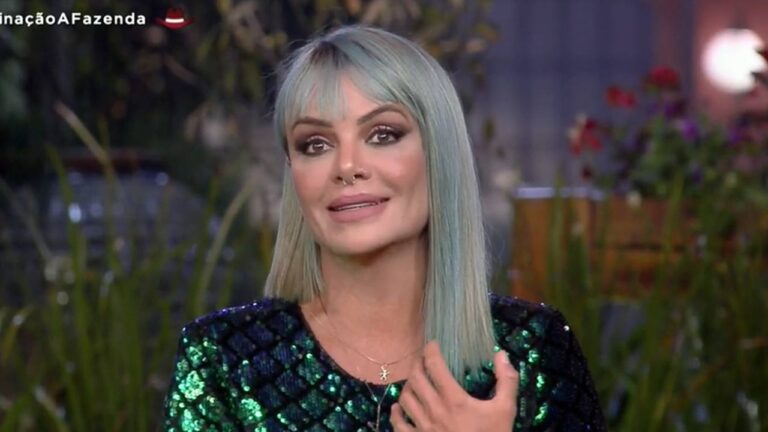 Mulher com cabelo liso azul-claro e franja fala diante da câmera; ela usa vestido brilhante em tons de verde e azul e aparece em um cenário noturno com plantas desfocadas ao fundo.