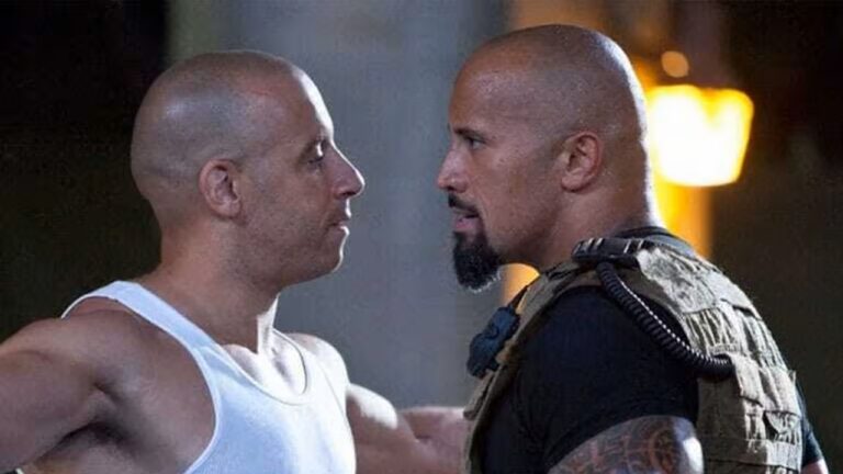 Dois homens carecas e musculosos aparecem frente a frente, muito próximos, em um confronto tenso. O homem à esquerda usa uma camiseta branca sem mangas; o da direita veste roupa escura com um colete tático e tem barba curta. Ambos mantêm expressões sérias e olhares fixos um no outro. O fundo é escuro, com iluminação quente desfocada, sugerindo uma cena de ação ou conflito intenso em um filme.