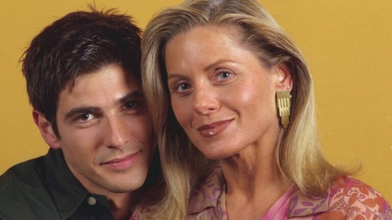 Homem de cabelo escuro e mulher loira posam próximos em retrato de estúdio com fundo amarelo e expressões suaves olhando para a câmera