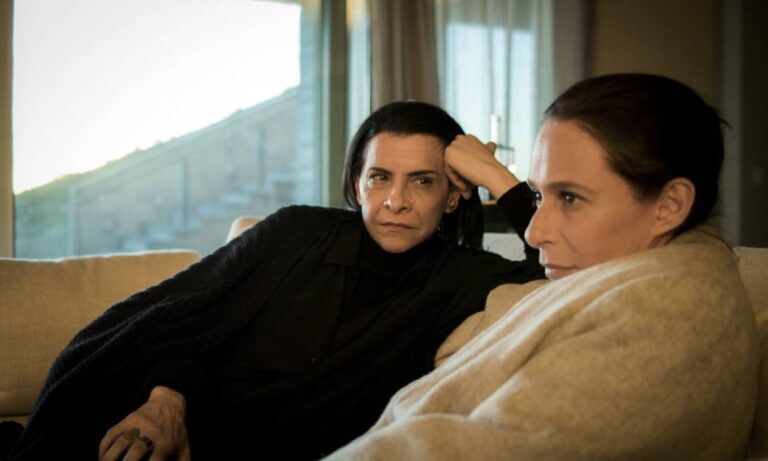 Duas mulheres sentadas em um sofá dentro de uma sala iluminada por luz natural. Ambas vestem roupas em tons escuros e mantêm expressões sérias e pensativas. Uma delas apoia o braço no encosto do sofá e observa a outra, que olha para a frente envolta em um cobertor claro. Ao fundo, há uma janela ampla com cortinas e uma paisagem externa desfocada.