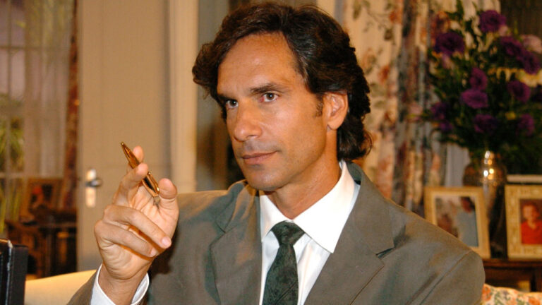 Victor Fasano como James Perkins na novela América segurando uma caneta dourada e usando terno cinza em cena