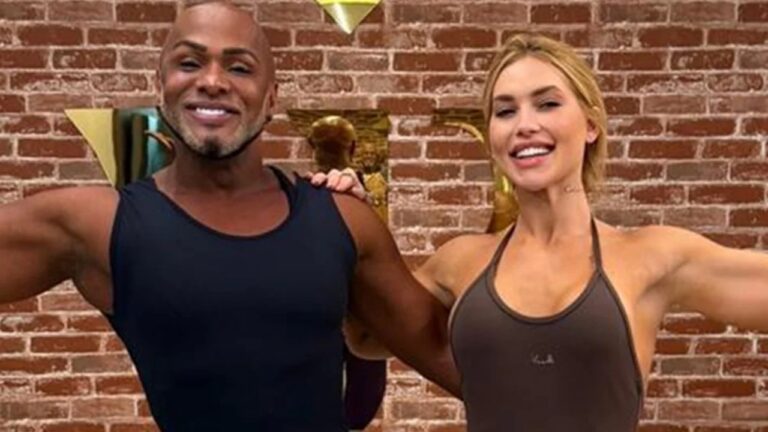 Duas pessoas posam sorrindo em frente a uma parede de tijolos aparentes, ambas usando roupas esportivas. Elas aparecem lado a lado com os braços erguidos, demonstrando clima descontraído e energia de treino em ambiente de academia ou estúdio fitness.