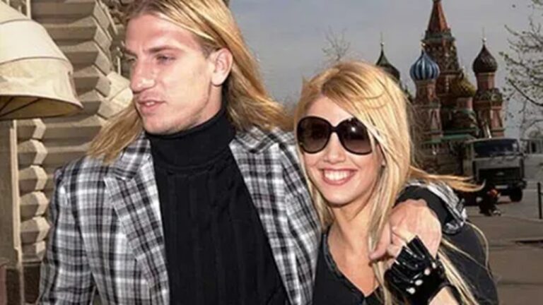 Casal posa sorridente enquanto caminha ao ar livre em uma área urbana. A mulher usa óculos escuros grandes e veste roupa preta; o homem, de cabelos longos e loiros, usa um casaco xadrez e gola alta. Ao fundo, aparecem construções de arquitetura russa, incluindo cúpulas coloridas características, sob céu nublado.