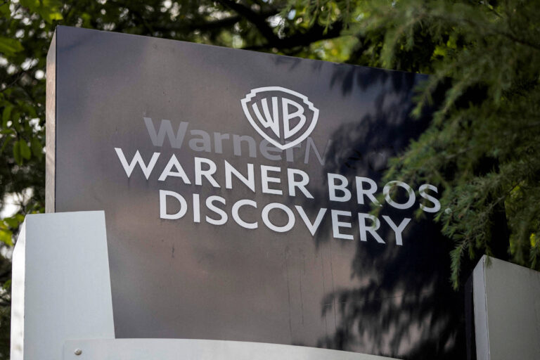 Placa com logotipo e nome da Warner Bros Discovery cercada por árvores na entrada da sede da empresa
