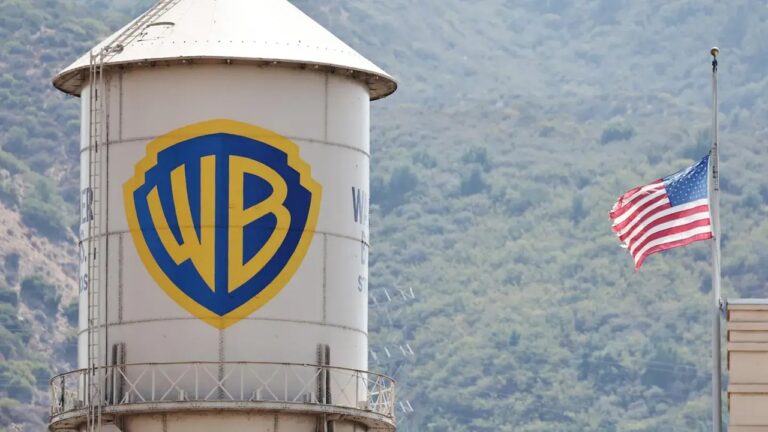 Caixa d’água com logotipo da Warner Bros ao lado de bandeira dos Estados Unidos em mastro diante de colina arborizada