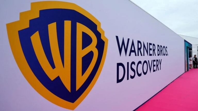 Parede branca de evento com o logotipo da Warner Bros. Discovery em destaque: escudo azul e dourado da Warner Bros. à esquerda e o nome “Warner Bros. Discovery” em letras pretas à direita. Um tapete rosa percorre o chão ao longo da parede, sugerindo um ambiente de evento ou tapete vermelho.