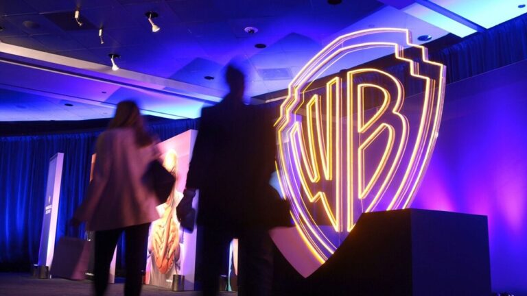 Um grande logotipo iluminado da Warner Bros. (WB) domina o cenário de um evento interno, com luzes em tons de azul e roxo. Duas pessoas aparecem em primeiro plano caminhando, vistas em silhueta e levemente desfocadas, sugerindo movimento. O ambiente lembra uma apresentação corporativa ou convenção, com palco, painéis e iluminação cênica profissional.