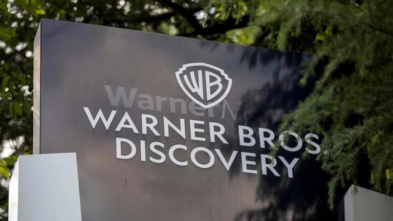 A imagem mostra uma placa da Warner Bros. Discovery, com o logotipo “WB” no topo e o nome da empresa escrito em letras brancas sobre um fundo escuro. A placa está posicionada em um ambiente externo, cercada por árvores e vegetação.