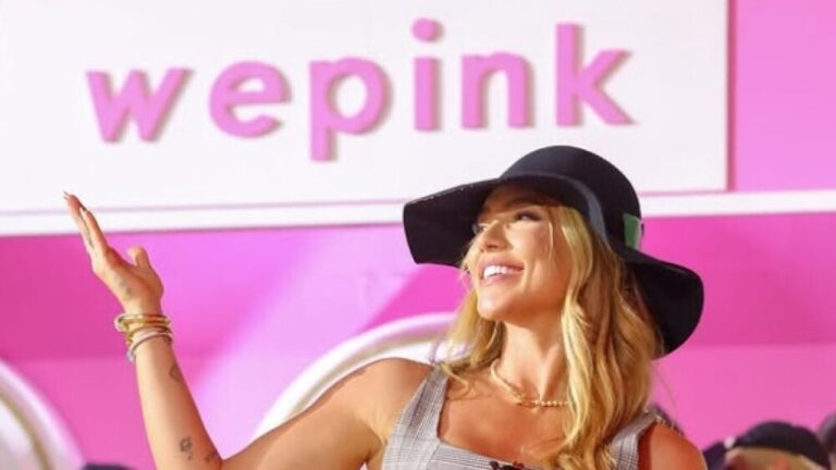 Mulher sorridente com chapéu preto e cabelos loiros soltos levanta uma das mãos em gesto descontraído em frente a um painel rosa e branco com o logotipo “wepink” em letras grandes cor-de-rosa