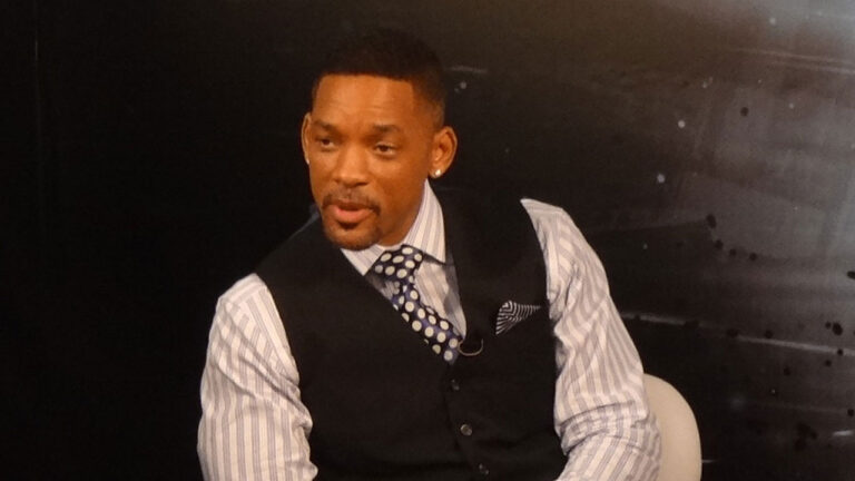 Will Smith veste colete preto e gravata de bolinhas sobre camisa listrada enquanto participa de entrevista em estúdio de televisão
