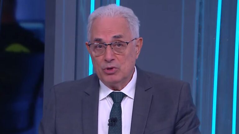 A imagem mostra William Waack, um homem mais velho, de cabelos brancos, usando terno cinza, camisa branca e gravata verde escura. Ele também usa óculos e está falando em frente a um fundo azul com luzes verticais, no estúdio da CNN Brasil