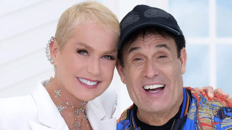 Xuxa Meneghel de blazer branco e Sérgio Mallandro de boné preto sorriem abraçados com fundo de janela branca