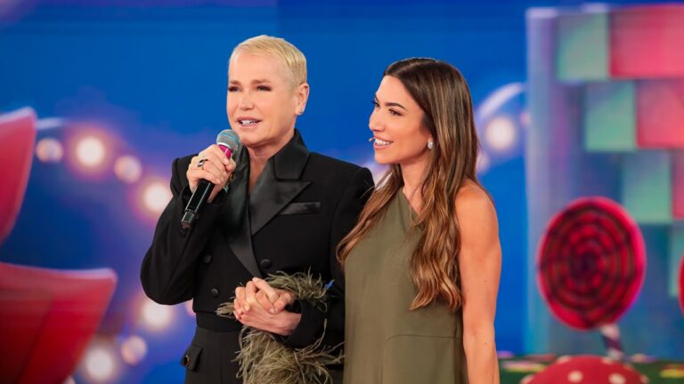 Duas mulheres em um palco colorido seguram as mãos e sorriem, uma delas com cabelo loiro curto veste terno preto e segura um microfone, enquanto a outra, de cabelo longo e castanho, usa vestido verde, ambas com microfones de lapela presas ao rosto diante de um fundo iluminado com luzes e elementos decorativos coloridos