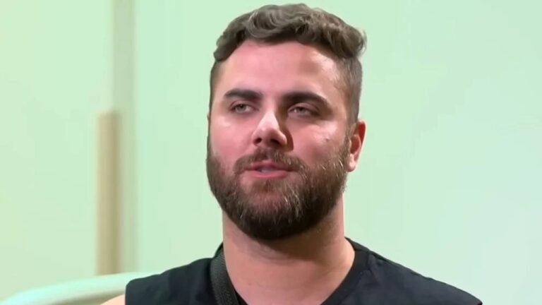 Homem de barba cheia e cabelos curtos e ondulados veste camiseta preta e tem expressão séria, em ambiente interno com parede clara desfocada ao fundo.