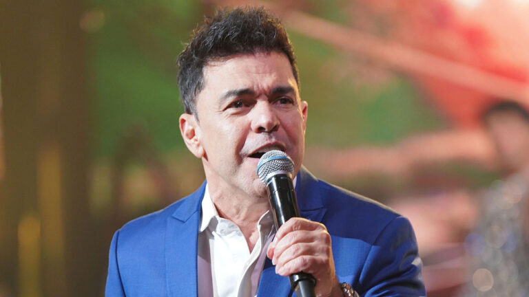 Zezé Di Camargo canta com microfone no palco do Caldeirão com Mion vestindo paletó azul e camisa branca nos estúdios da Globo
